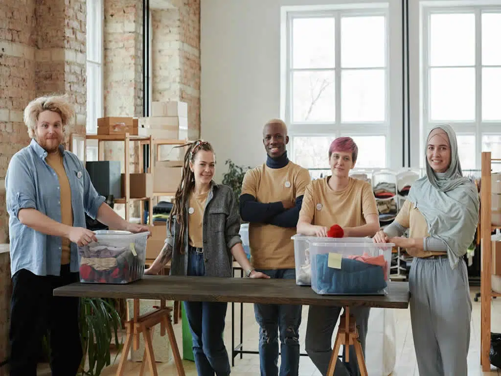 Group of diverse volunteers in casual attire standing behind a table with donation bins, showcasing community engagement and support for non-profit initiatives.