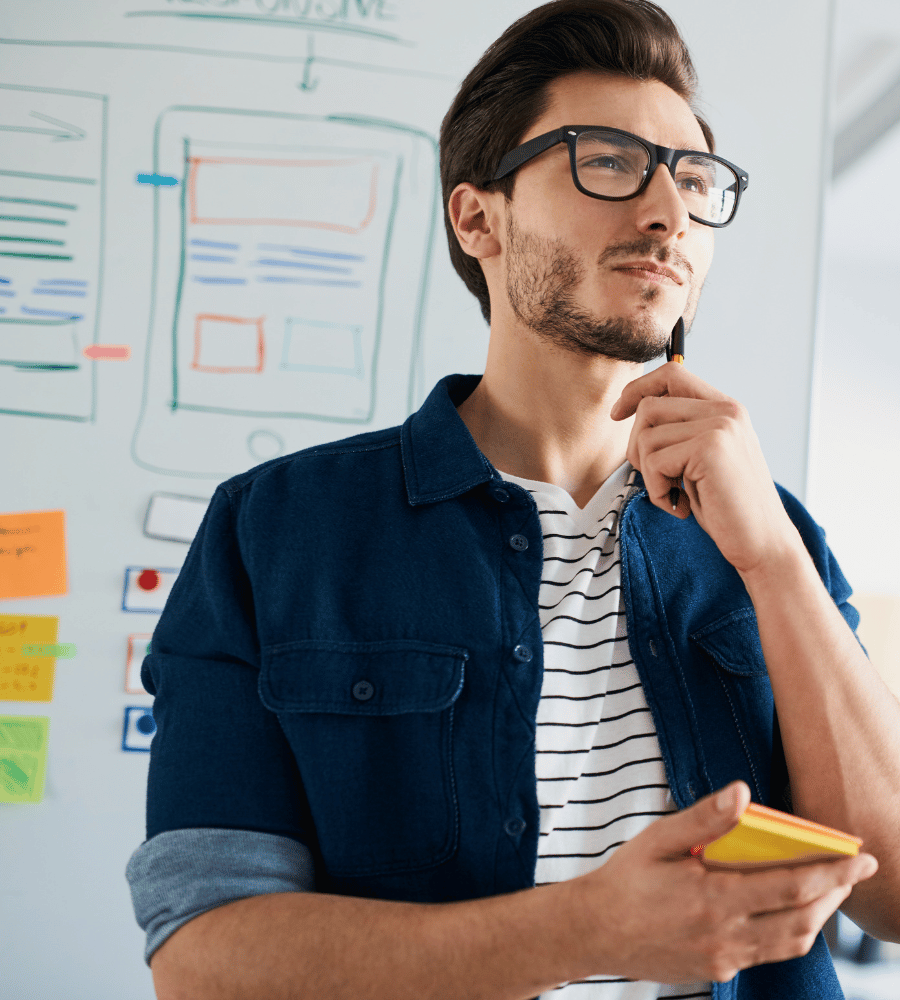 Man contemplating web design ideas while holding a notepad in front of a whiteboard with sketches and sticky notes, illustrating the creative process in digital development.